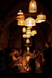 Candlelit intimate dinner at a long communal table beneath woven wicker pendant lamps in a stone-arched dining room, warm cozy ambiance