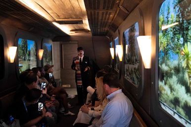 Passengers in a dimly lit tour train car listen to a guide as desert landscape glides past large windows; some people film the scene on phones.