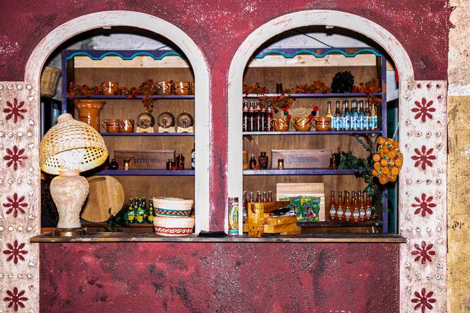 Colorful Mexican-style double-arched market window display with handmade clay pottery, stacked woven baskets, a wicker lamp, rows of bottled drinks and decorative garlands on wooden shelves against painted stucco.