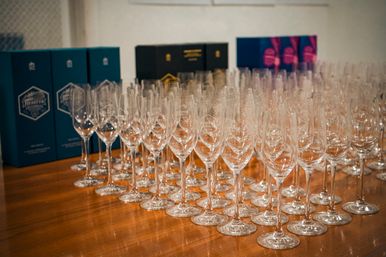 Neatly arranged rows of empty wine glasses on a polished wooden table, ready for a toast at a wine-tasting or reception, with teal and black boxes blurred in the background.