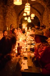 Cozy candlelit dinner in a stone-vaulted cellar — long communal table of diners under hanging wicker lamps, glowing candles and floral centerpieces in warm amber light.