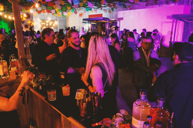 Indoor urban warehouse cocktail party with colorful string lights and papel picado banners, guests chatting at a crowded bar with cocktails and mixers on the counter