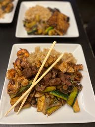 Sizzling teppanyaki-style plate with grilled steak, shrimp, zucchini and mushrooms over fried rice, crossed chopsticks on a white square plate.