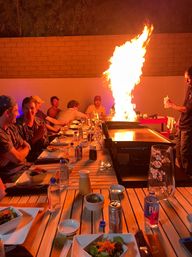 Evening backyard dinner party around a teppanyaki-style flat-top grill on a patio, chef creating a dramatic towering flame as guests sit at a long wooden table set with salads, bottles and drinks.