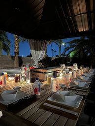 Inviting outdoor nighttime dining on a long wooden patio table under a covered pergola, warm fairy lights and candles, bottled water and white dishware neatly arranged, palm trees silhouetted against a deep-blue twilight sky.