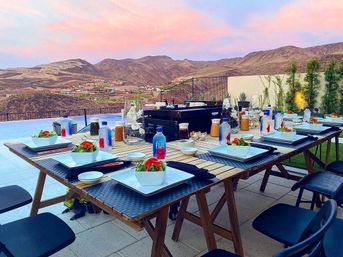 Poolside patio dining at sunset with a long wooden table set with salads, bottled water, condiments and candles, framed by a pink sky and rugged desert mountain view
