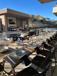 Poolside outdoor dining in a modern backyard on a sunny day — long wooden table set with white plates and bowls, bottled water, black napkins, folding chairs, grill station and covered patio lounge.