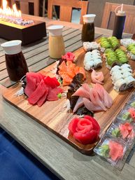 Vibrant sushi platter on a wooden board with tuna, salmon and yellowtail sashimi, avocado-topped and rice rolls, pickled ginger and wasabi packets, condiment bottles on a patio table beside a tabletop fire feature.