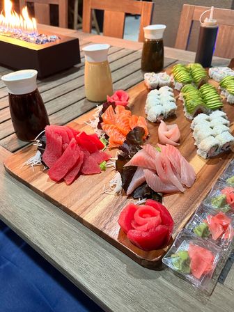 Vibrant sushi platter on a wooden board with tuna, salmon and yellowtail sashimi, avocado-topped and rice rolls, pickled ginger and wasabi packets, condiment bottles on a patio table beside a tabletop fire feature.
