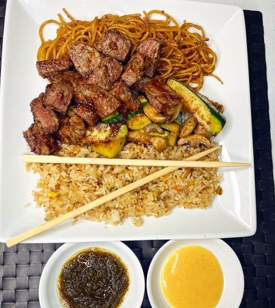 Mouthwatering grilled steak cubes with fried rice, stir-fried noodles and sautéed zucchini and mushrooms on a square white plate, chopsticks laid across the rice, with two small bowls of green herb sauce and creamy orange dipping sauce on a woven placemat.