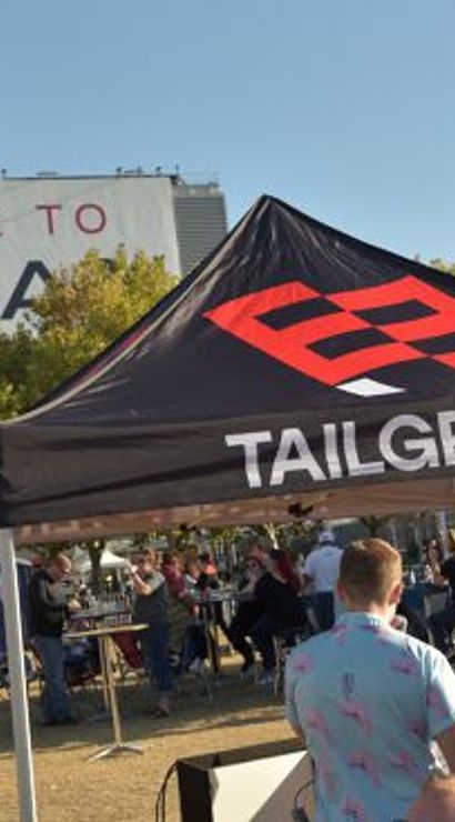 Tailgate scene outside a football stadium with a black canopy featuring a red logo and white lettering, fans mingling, portable toilets, and a large stadium welcome sign under a clear blue sky.
