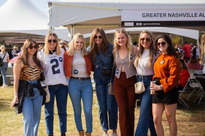 Seven women in casual fall outfits smiling and posing at an outdoor Nashville tailgate event under white tents, wearing denim, sunglasses and holding drinks