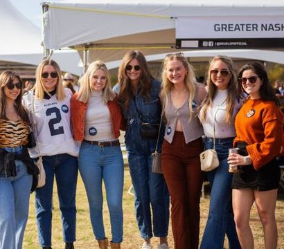 Seven women in casual fall outfits smiling and posing at an outdoor Nashville tailgate event under white tents, wearing denim, sunglasses and holding drinks