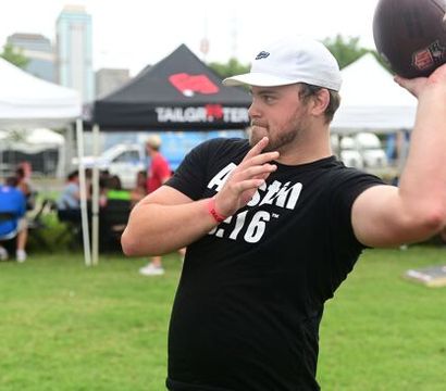 Young man in a white cap and black T‑shirt throwing a football at an outdoor tailgate on a grassy field with vendor tents and people in the background.