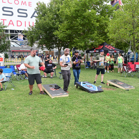 Adults playing cornhole at a stadium tailgate on a grassy lawn, tossing bean bags between wooden boards with folding chairs, vendor tent, and trees in the background.