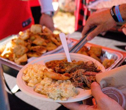 Heaping paper plate of comfort food - mac and cheese, potato salad, pulled pork and fried appetizers - being served at an outdoor barbecue or food festival.