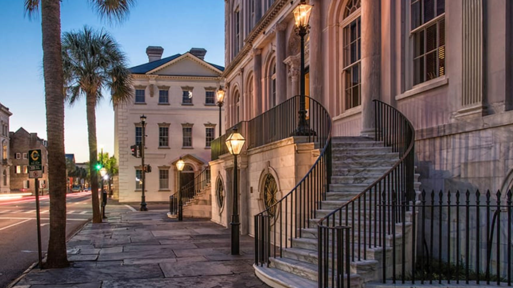 Historic southern streetscape at dusk featuring a curved marble staircase, iron railings, palm trees and glowing gaslamp-style streetlights.