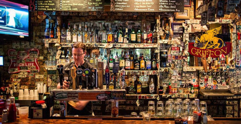 Cozy downtown dive bar interior with a bartender pouring draft beer, multiple taps and liquor bottles, walls plastered with dollar bills and stickers, a chalkboard beer menu and glassware on a wooden bar.