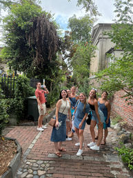 Group of friends smiling and posing on a tree-lined historic brick and cobblestone alley beside a red brick wall and lush greenery during a casual summer stroll.