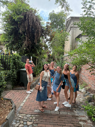 Group of friends smiling and posing on a tree-lined historic brick and cobblestone alley beside a red brick wall and lush greenery during a casual summer stroll.