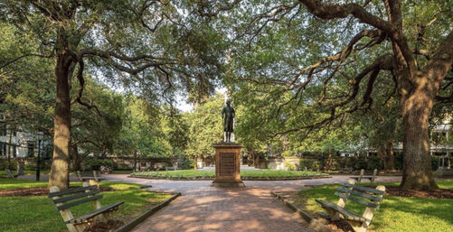 Bronze statue on a stone pedestal centered in a circular, oak-shaded city park with sprawling live oaks, brick walkway, manicured flowerbed and wooden benches.