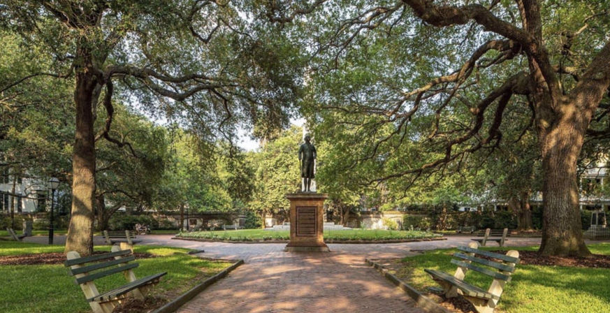 Bronze statue on a stone pedestal centered in a circular, oak-shaded city park with sprawling live oaks, brick walkway, manicured flowerbed and wooden benches.