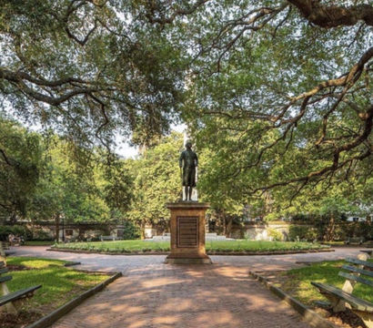 Bronze statue on a stone pedestal centered in a circular, oak-shaded city park with sprawling live oaks, brick walkway, manicured flowerbed and wooden benches.