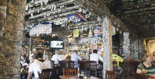 Lively bar interior with patrons at wooden tables and a long bar, ceiling and walls plastered with U.S. dollar bills and notes, TV above stocked liquor shelves