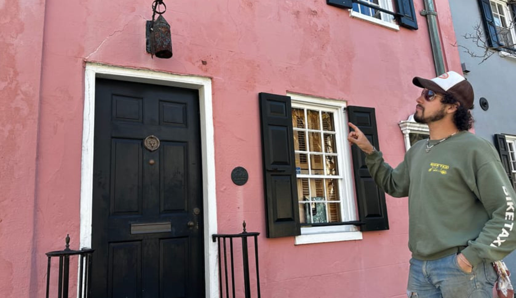 Person in a cap and sunglasses pointing at a pastel pink townhouse facade featuring a black door, black shutters, white-trimmed window and small iron railing.