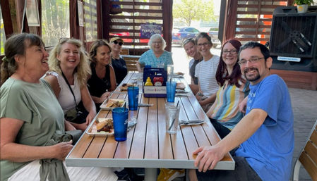 Ten smiling adults sharing a casual outdoor patio meal at a long wooden table with plates of barbecue, blue drinking cups, condiments, and sunlight filtering through slatted walls.