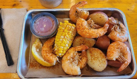Cajun-style shrimp boil in a metal tray with seasoned whole shrimp, red potatoes, corn on the cob, lemon wedge and dipping sauce on a wooden table