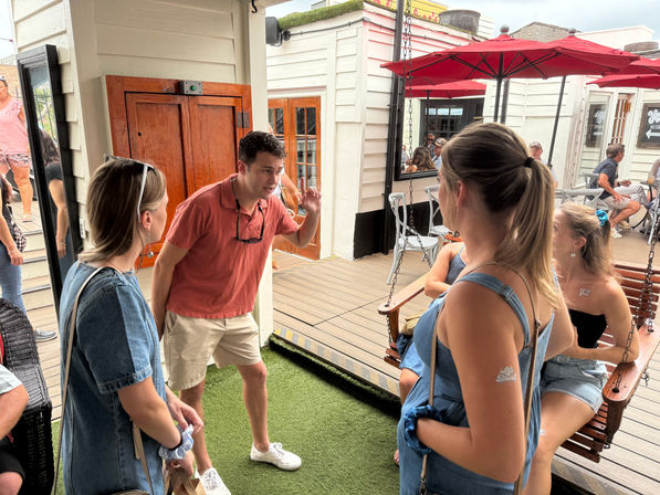 Man animatedly talking to friends on a lively outdoor café patio with red umbrellas, wooden swing and deck seating