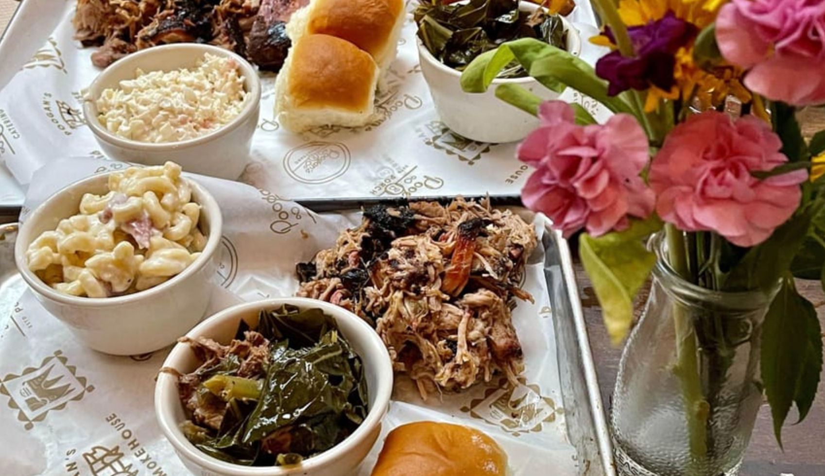 Southern BBQ tray with pulled pork, collard greens, creamy macaroni salad, coleslaw and slider buns on parchment-lined metal trays, next to a jar of pink flowers