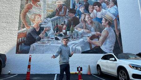 Person posing in a parking lot in front of a large colorful outdoor mural of people dining on a white brick wall, with parked cars and orange traffic cones nearby.