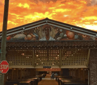 Brick open-air market pavilion on State Street with wooden vendor stalls, a colorful mural of a figure and produce above the entrance, a stop sign in the foreground and a fiery orange sunset sky.