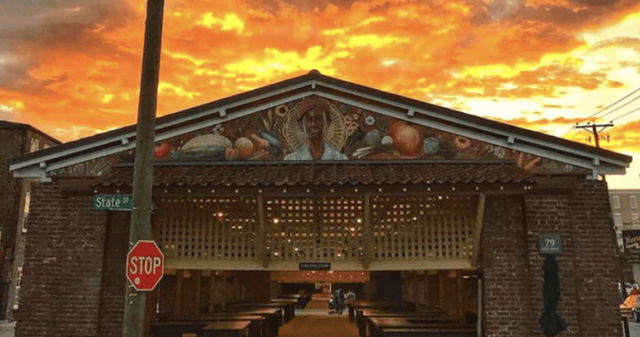 Brick open-air market pavilion on State Street with wooden vendor stalls, a colorful mural of a figure and produce above the entrance, a stop sign in the foreground and a fiery orange sunset sky.