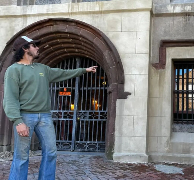 Person in green sweatshirt and cap pointing at a barred stone-arch entrance with a 'No Trespassing' sign, brick sidewalk outside a historic urban building.