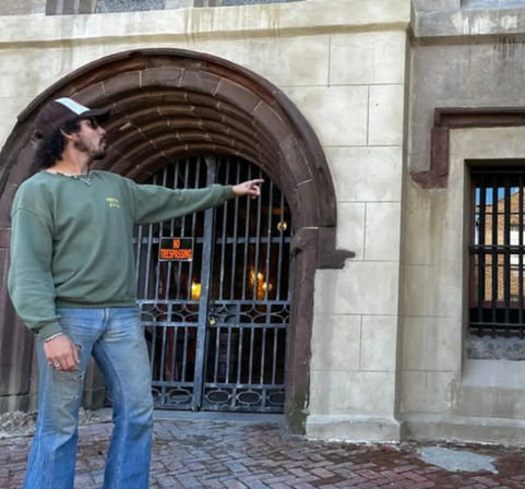 Person in green sweatshirt and cap pointing at a barred stone-arch entrance with a 'No Trespassing' sign, brick sidewalk outside a historic urban building.