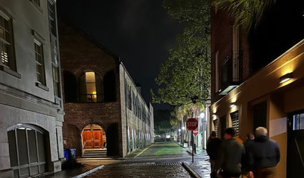 Nighttime historic downtown scene: wet cobblestone alley flanked by brick buildings with a warm-lit arched doorway, tree-lined sidewalk, stop sign, and a small group of people walking under streetlights.