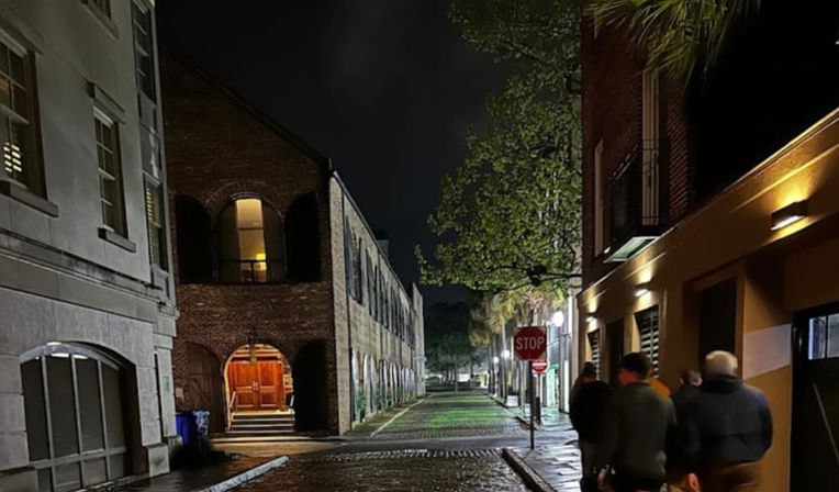 Nighttime historic downtown scene: wet cobblestone alley flanked by brick buildings with a warm-lit arched doorway, tree-lined sidewalk, stop sign, and a small group of people walking under streetlights.