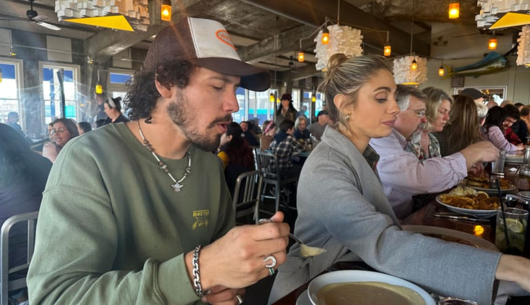 Crowded casual restaurant bar with warm pendant lights; a man in a trucker hat and green sweatshirt scoops soup while a woman in a gray coat reaches for a plate among other diners.