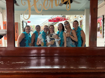 Seven friends wearing bright blue feather boas taking a mirror selfie and holding cocktails on a lively outdoor bar patio under an "Est. 1930" sign.