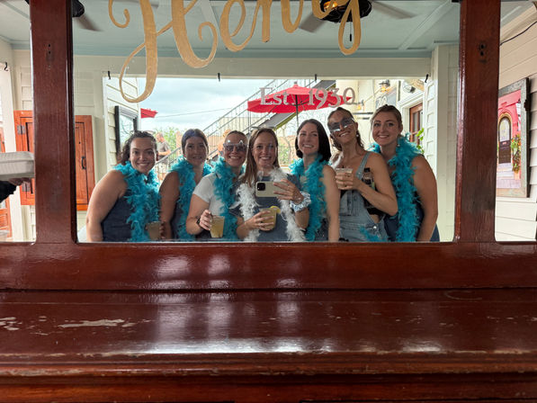 Seven friends wearing bright blue feather boas taking a mirror selfie and holding cocktails on a lively outdoor bar patio under an "Est. 1930" sign.