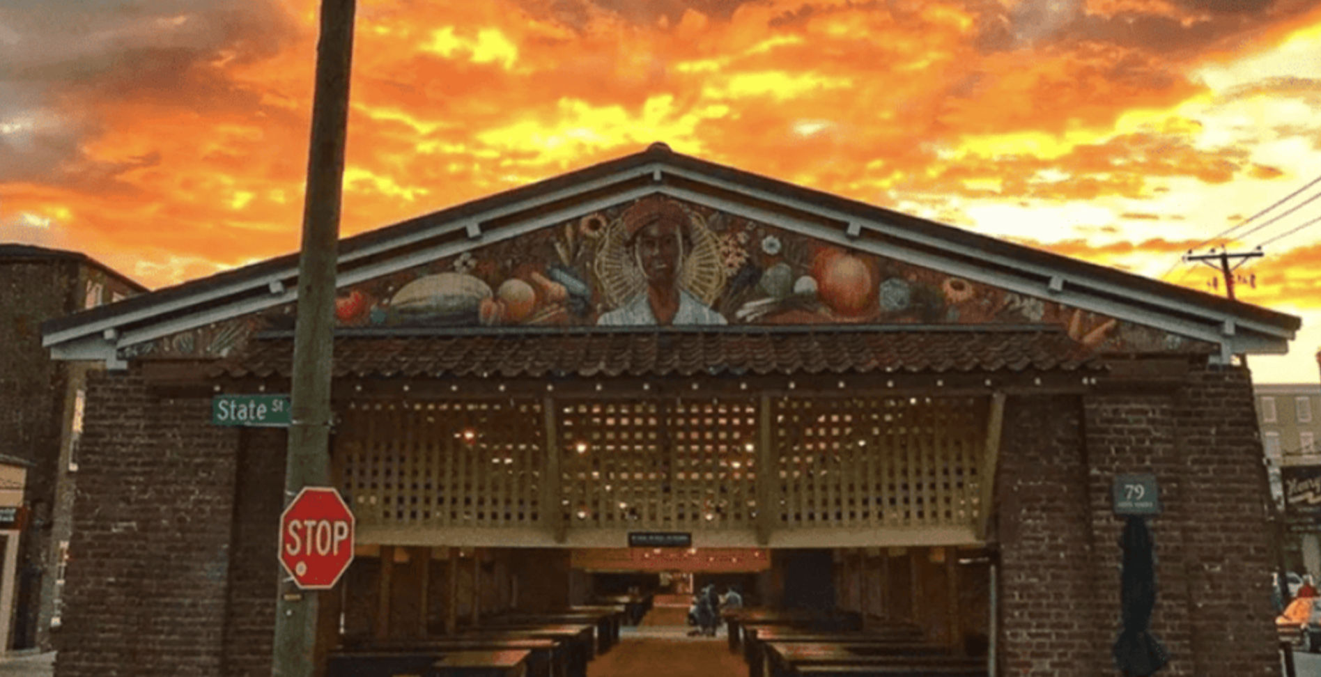 Open-air brick market pavilion on State Street with a mural of a farmer and produce, empty wooden stalls, a stop sign in the foreground and a fiery orange sunset sky.