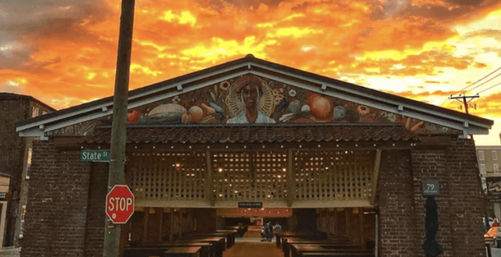 Open-air brick market pavilion on State Street with a mural of a farmer and produce, empty wooden stalls, a stop sign in the foreground and a fiery orange sunset sky.