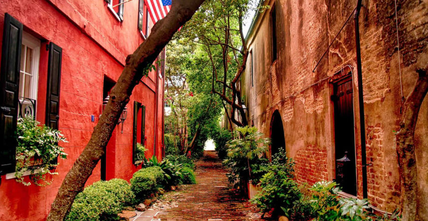 Quaint historic brick alley flanked by red stucco and weathered brick buildings, lush greenery and potted shrubs with an American flag overhead