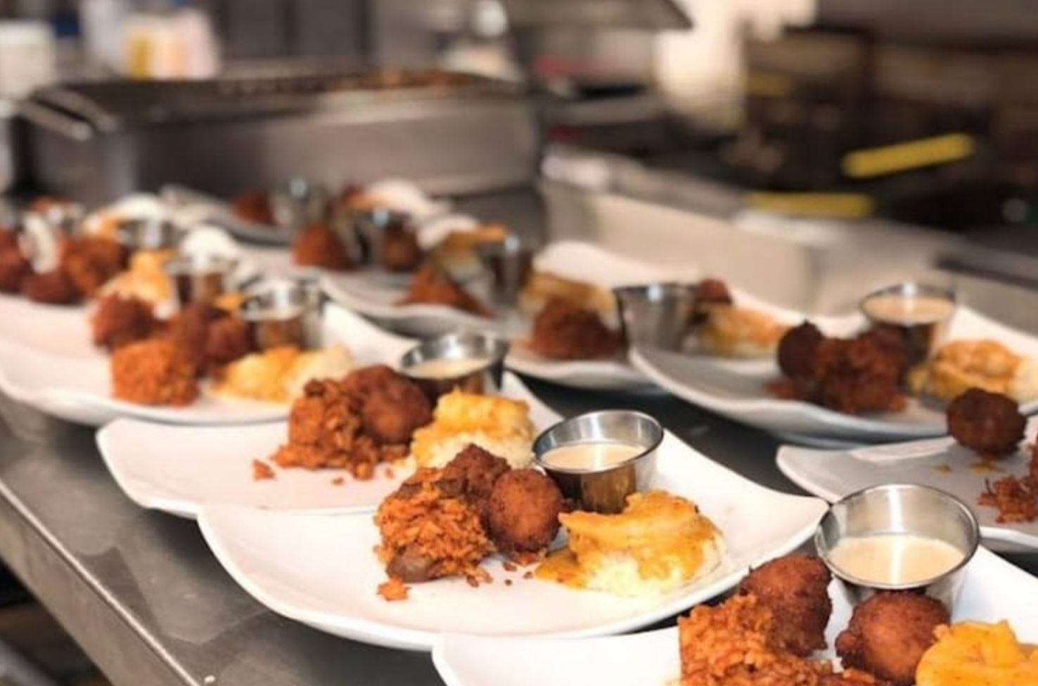Rows of white plates on a restaurant kitchen line with crispy fried chicken bites, cheesy mashed potatoes and small metal cups of dipping sauce.
