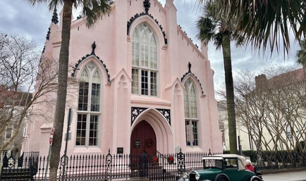 Pink Gothic Revival church with tall arched windows and black trim, framed by palm trees and a wrought-iron fence, a vintage green car parked on a cloudy historic downtown street.