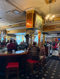 Lively vintage downtown pub interior with curved wooden bar, red-cushioned stools, patrons chatting, bartender at taps, hanging chandelier and framed artwork, patterned tile floor and warm daylight through front windows.