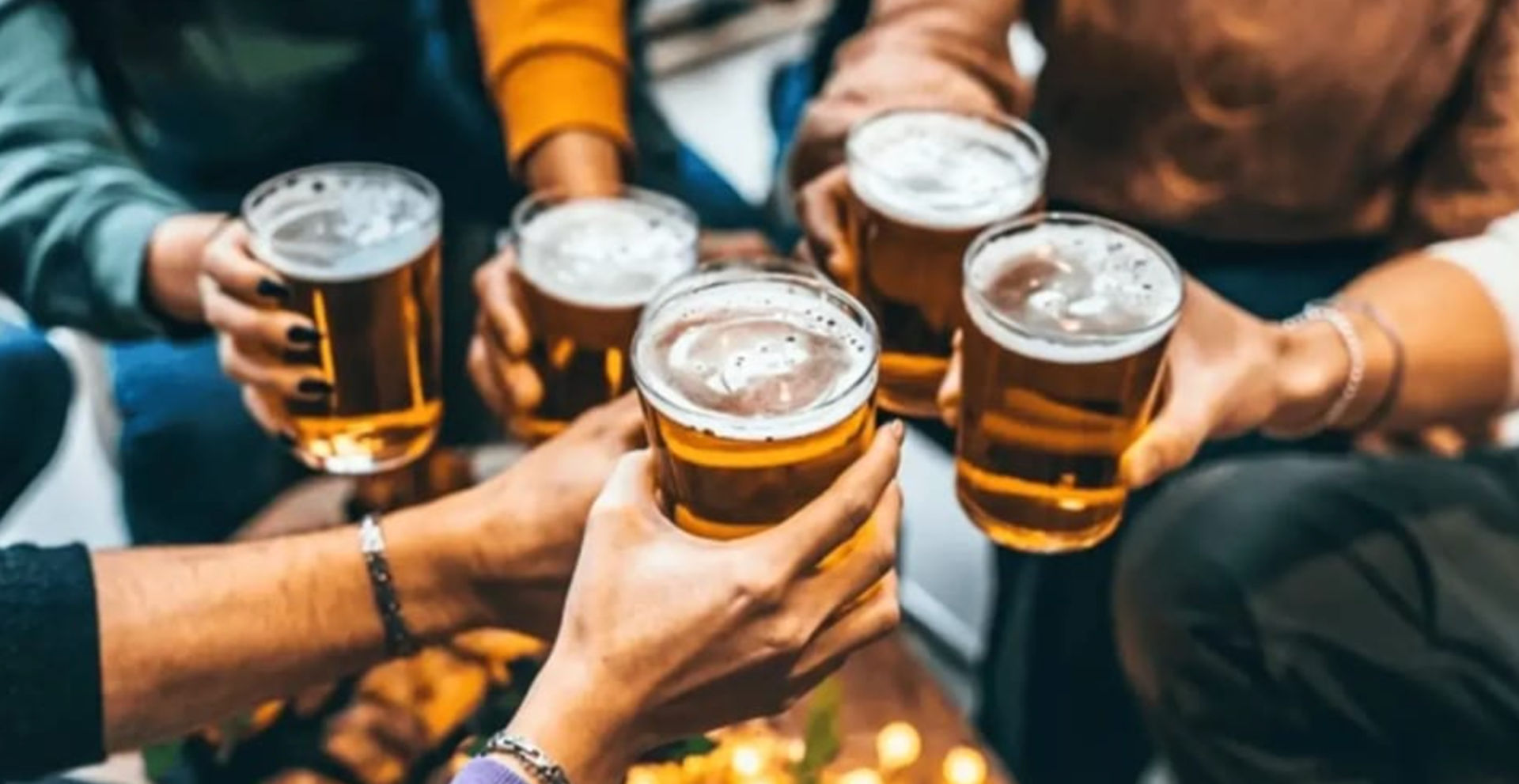 Close-up of several hands toasting pint glasses of amber beer, casual friends clinking drinks in a cozy bar or happy hour setting